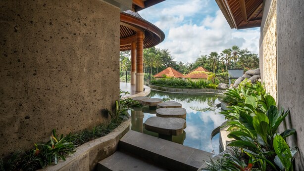 Uma área de piscina tranquila com um caminho de pedra que leva a um espaço coberto para sentar, cercada por vegetação exuberante e edifícios em estilo tradicional ao fundo.