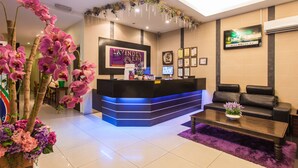 A hotel reception area with a black reception desk, purple flowers, a black leather sofa, and a TV mounted on the wall. - Lavender Inn Permas Jaya (Masai)