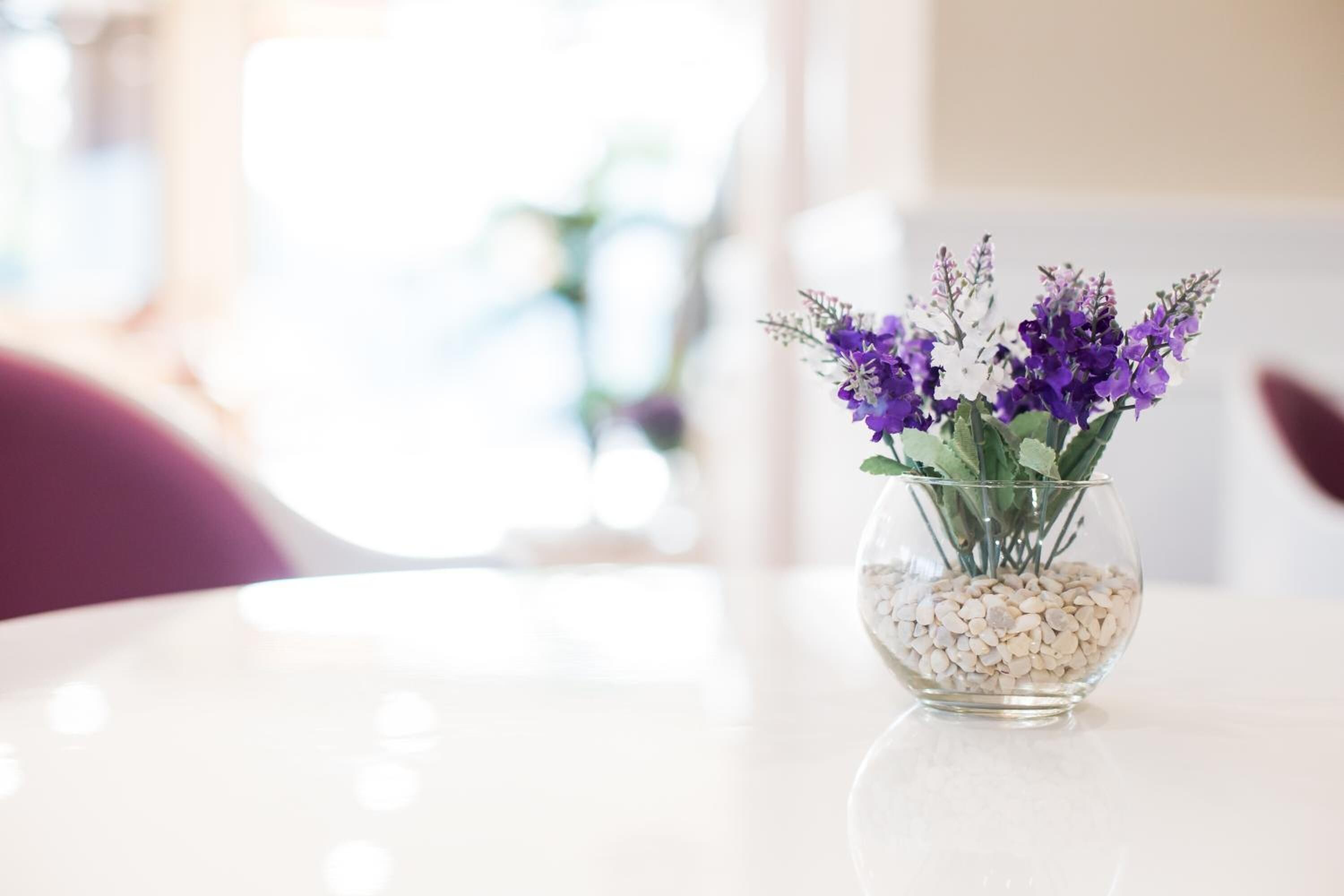 Eine klare Glasvase mit weißen Kieselsteinen und lila Blumen auf einem weißen Tisch.