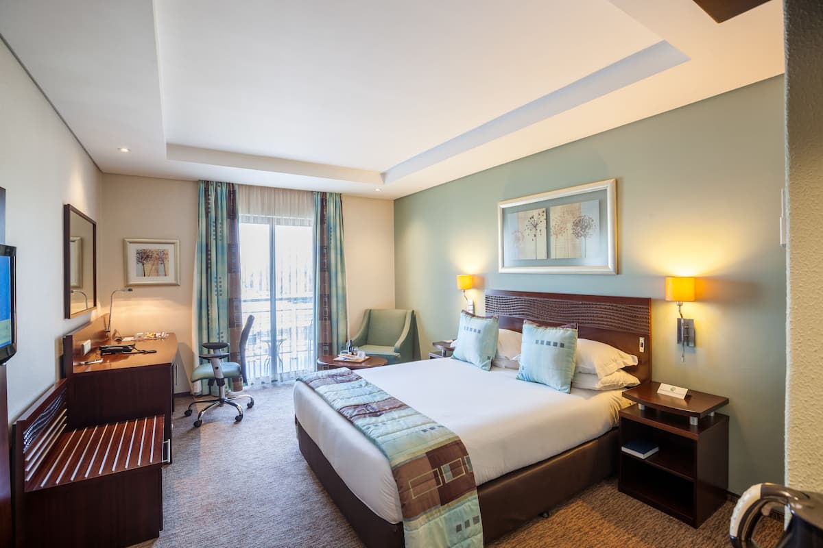 A hotel room with a large bed, a desk with a chair, a desk lamp, a chair, a bedside table with a lamp, and a framed picture on the wall.