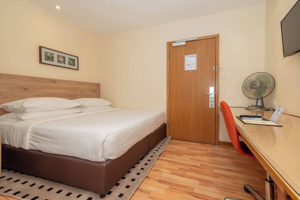 Een hotelkamer met een bed, een bureau met stoel, een houten deur en een ventilator aan de muur.