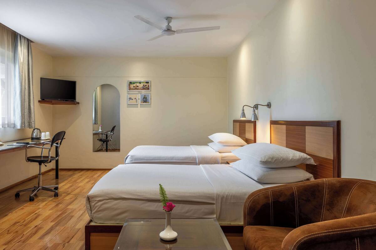 Hotelkamer met twee bedden, een bureau met stoel, een aan de muur bevestigde televisie en een plafondventilator.