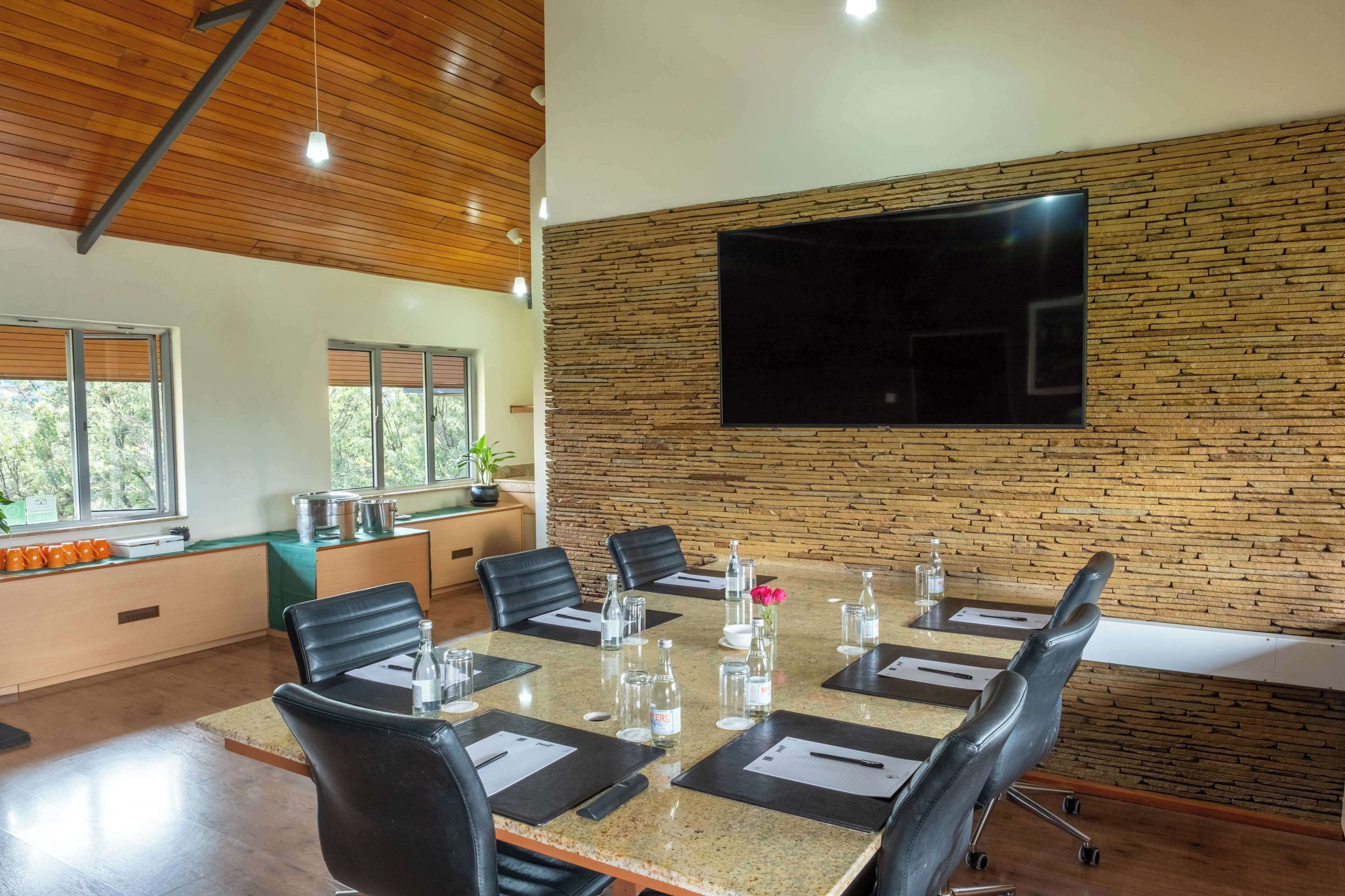 A conference room with a large table, black chairs, a flat-screen TV, and a brick-patterned wall.