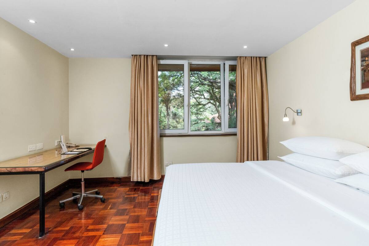 Habitación de hotel con piso de madera, un escritorio con silla, cama con ropa blanca y una ventana con cortinas, además de una lámpara fijada a la pared.