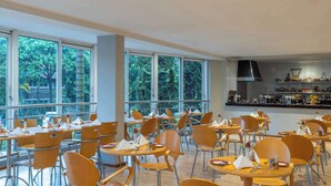 Un comedor amplio con varias mesas y sillas, ventanas grandes que ofrecen vista a árboles, y una cocina con electrodomésticos modernos.
