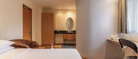 Quarto de hotel com cama, escrivaninha, cadeira, espelho e banheiro com pia e vaso sanitário.