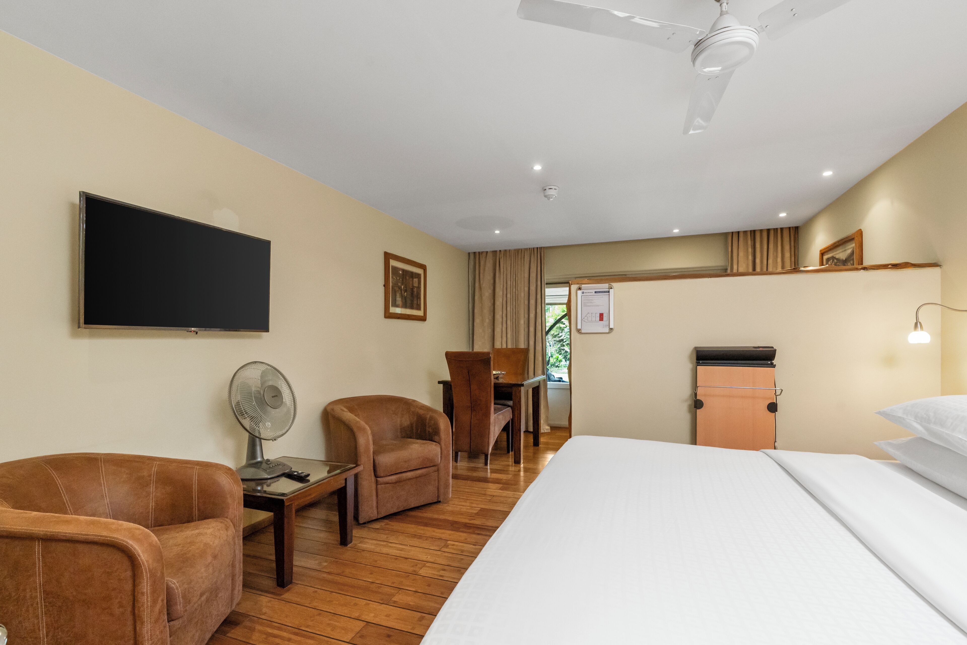 A hotel room with a bed, two armchairs, a desk, a flat-screen TV, a ceiling fan, and a small kitchenette.