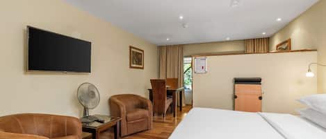 Une chambre d’hôtel comprenant un lit, deux fauteuils, un bureau, un téléviseur à écran plat, un ventilateur de plafond et une petite kitchenette.