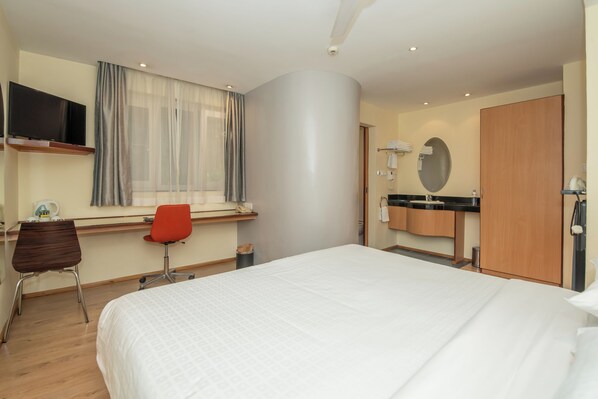 Habitación de hotel con cama, escritorio con silla, televisor, ventana con cortinas, baño con espejo y armario.