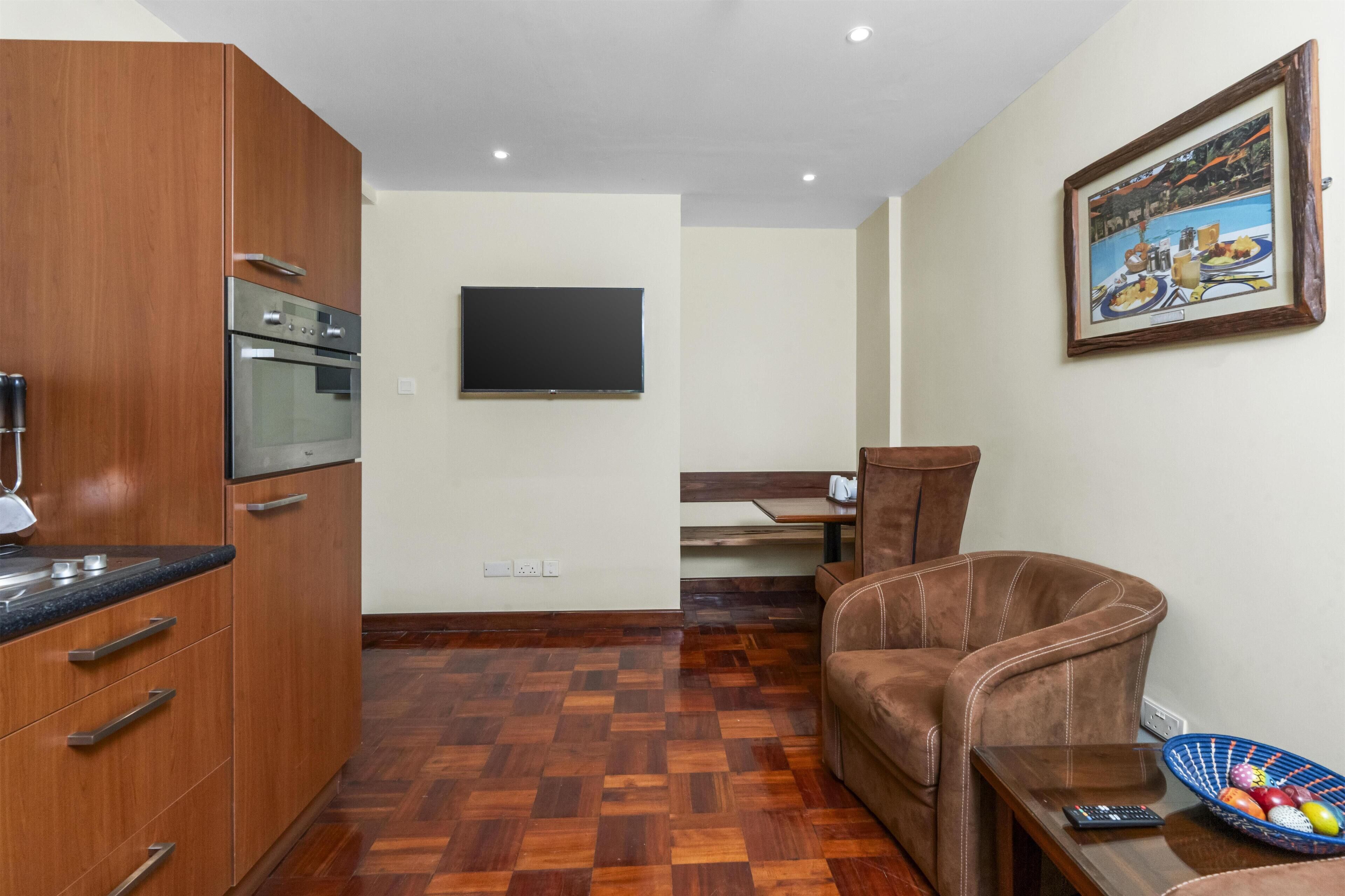 Un espacio habitable compacto con kitchenette, televisor de pantalla plana montado en la pared, sofá de cuero marrón y mesa de comedor con frutero azul.