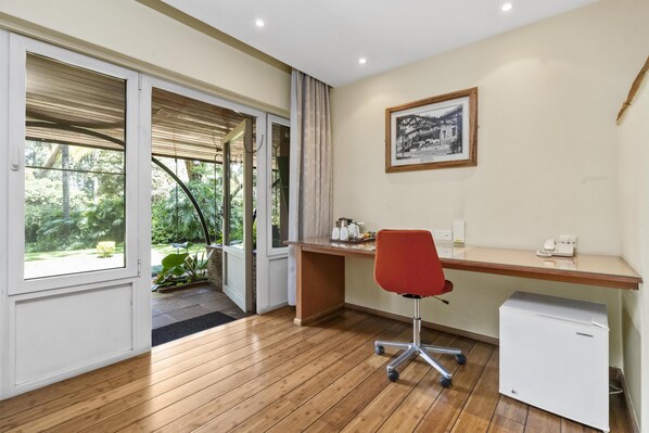 A room with a wooden floor, a desk with a chair, a small refrigerator, and a door leading to a garden.