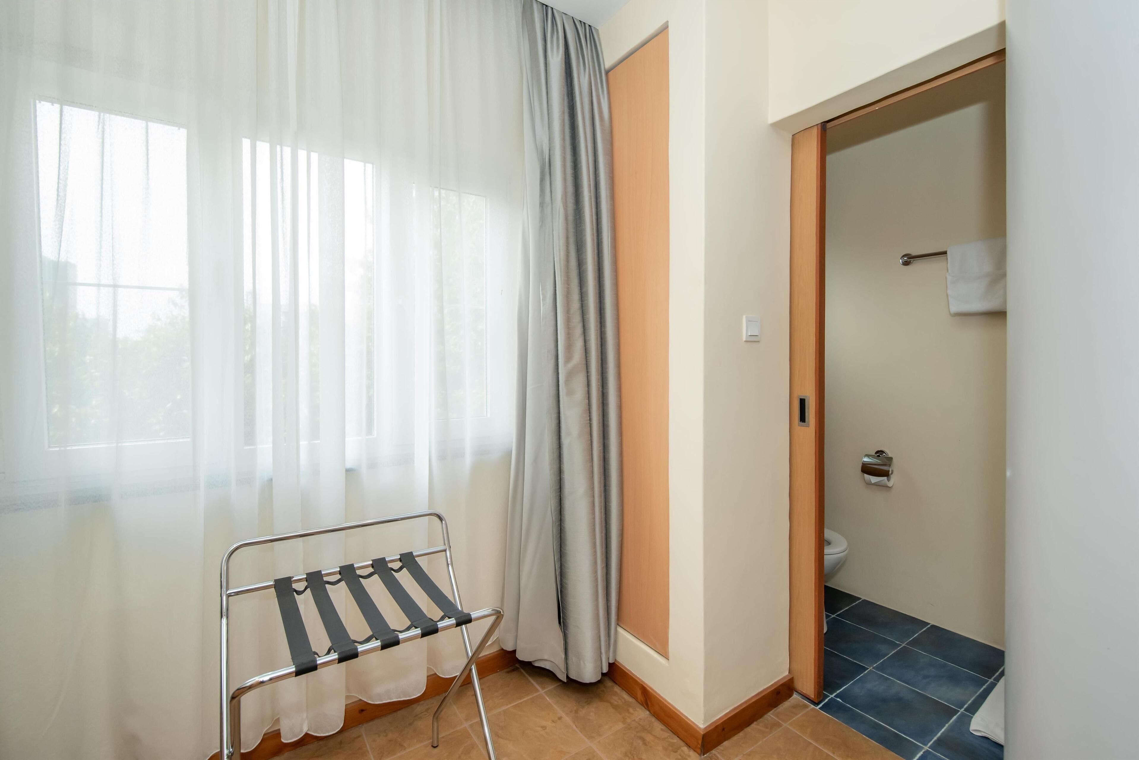 A room with a window covered by sheer curtains, a metal drying rack, a wooden door leading to a bathroom, and a tiled floor.