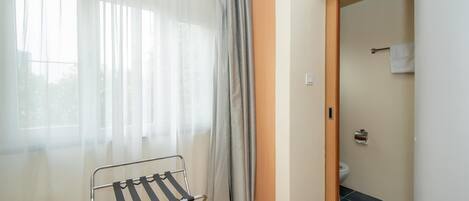A room with a window covered by sheer curtains, a metal drying rack, a wooden door leading to a bathroom, and a tiled floor.