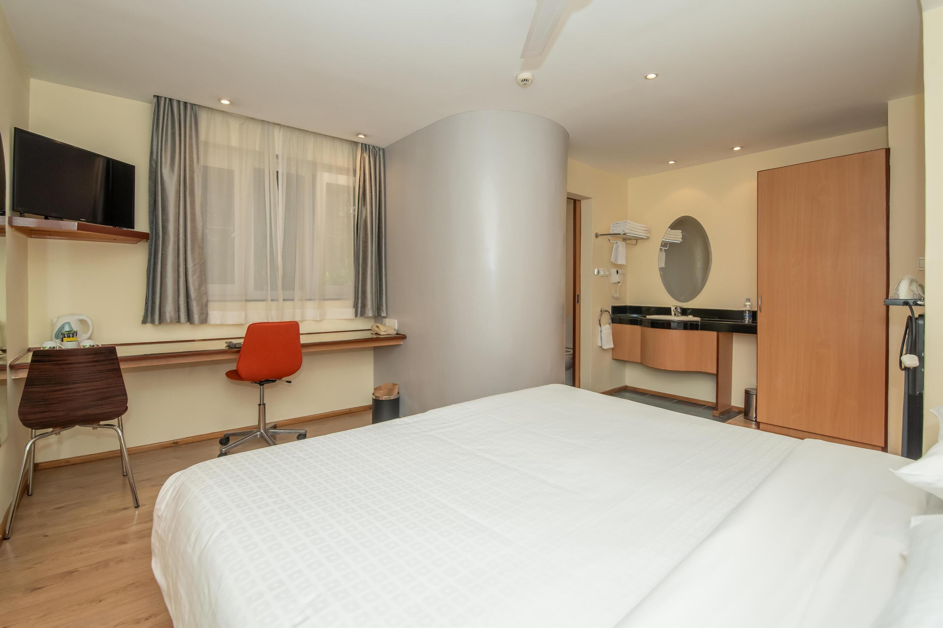 Habitación de hotel con cama, escritorio con silla, televisor montado en la pared, ventana con cortinas, baño con espejo y armario.