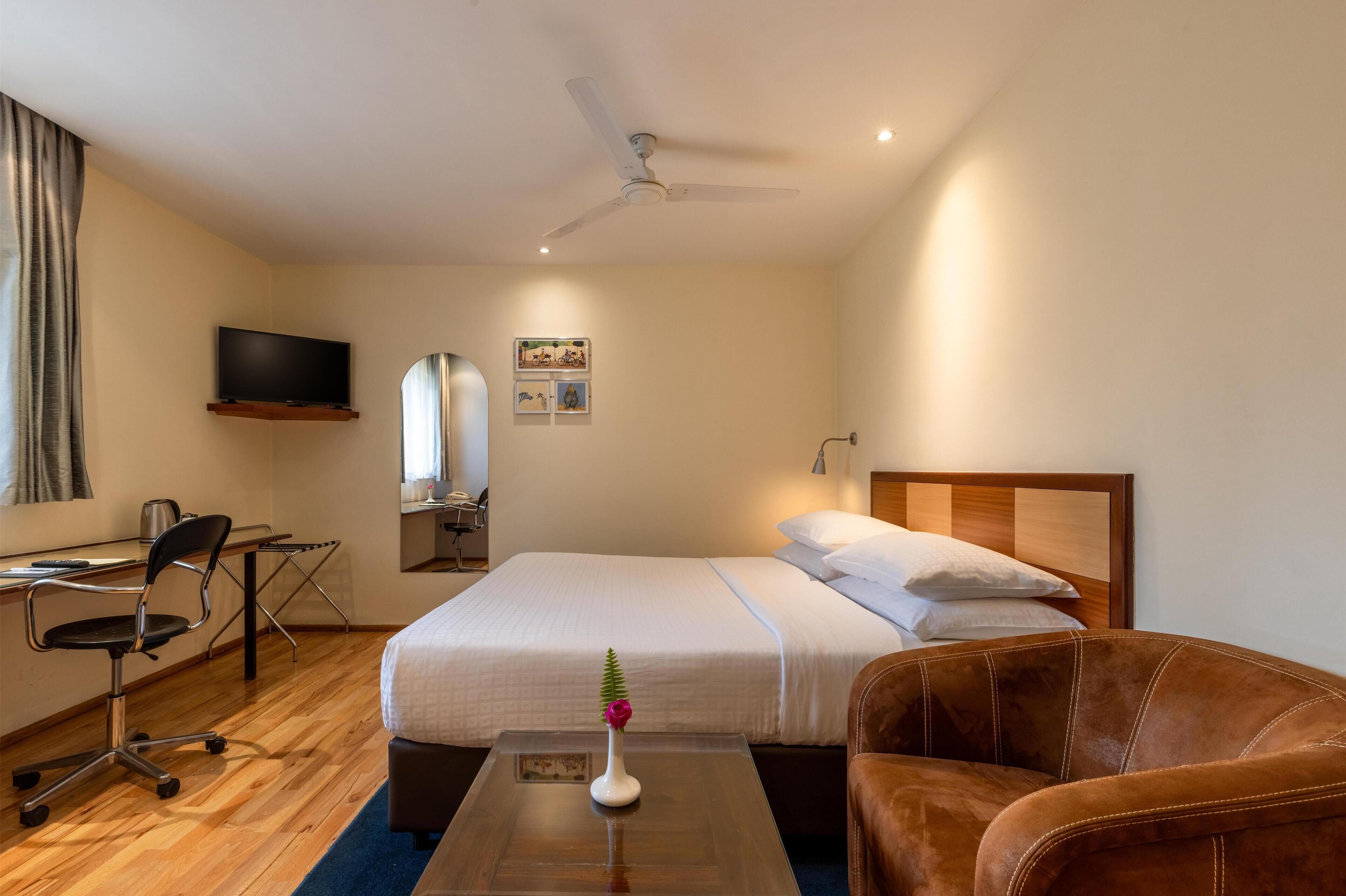 Ein Hotelzimmer mit einem Bett, einem Schreibtisch mit Stuhl, einem Fernseher, einem Deckenventilator und einem kleinen Tisch mit einer Vase.