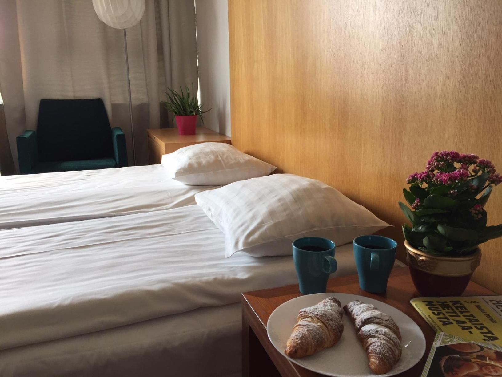 A hotel room with two beds, a wooden headboard, a small table with croissants, a potted plant, and a magazine.