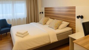 A neatly made bed with pillows, a chair, a bedside table, and a window with curtains. - Arkadia Hotel & Hostel (Helsinki)