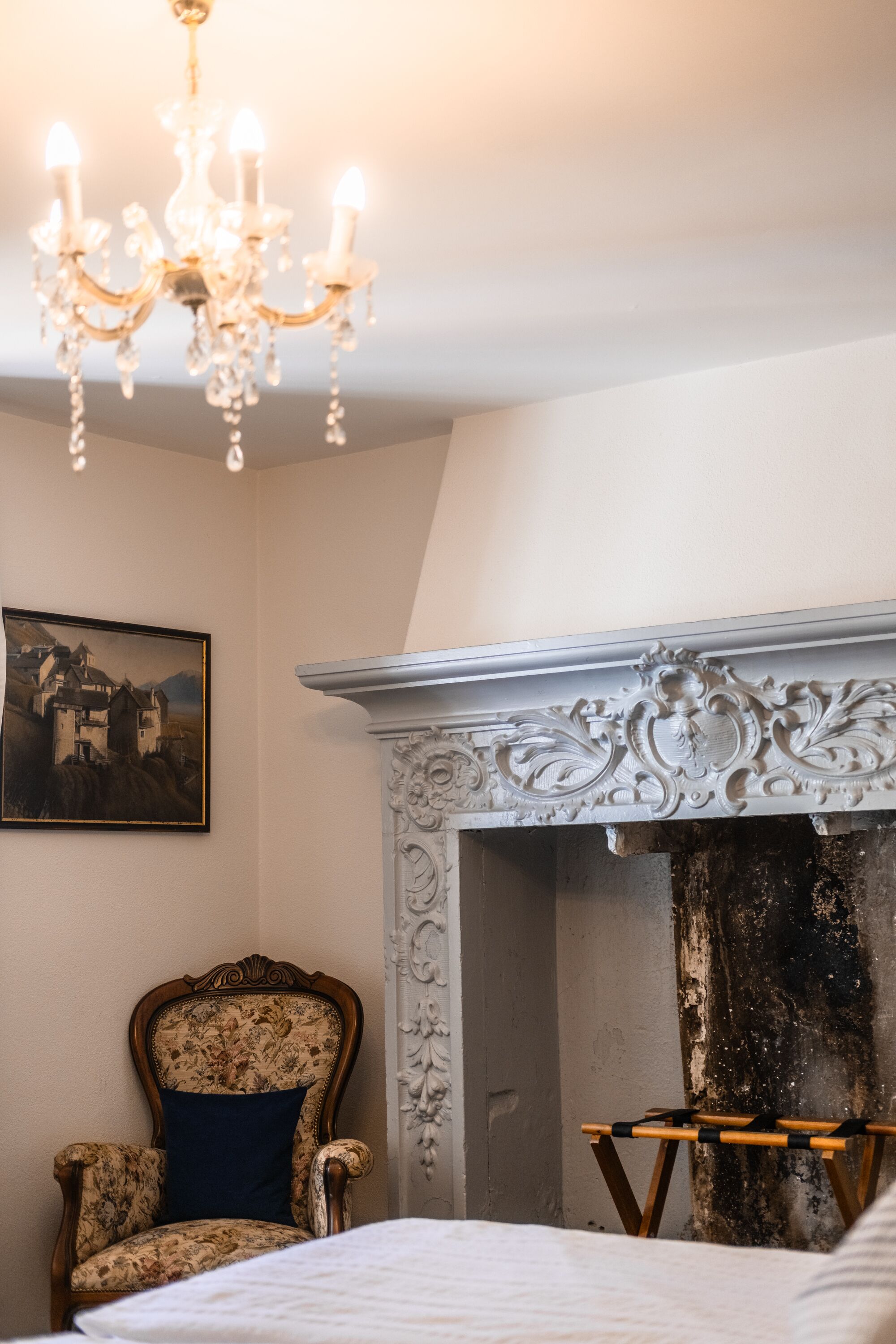 A bedroom with a bed, a chandelier, a fireplace, a chair, and a painting on the wall.