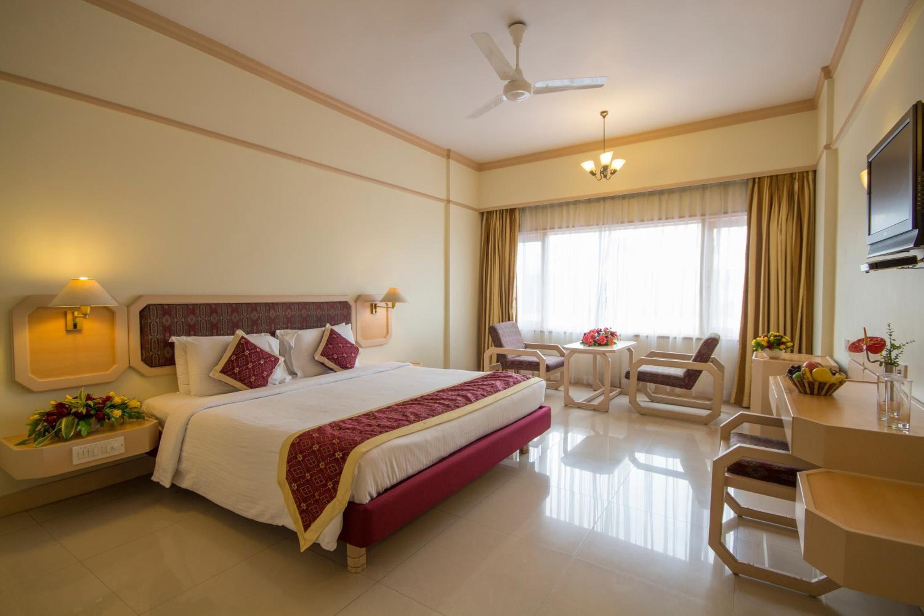 Une chambre d’hôtel avec un grand lit, un bureau avec une chaise, une télévision et un ventilateur de plafond.