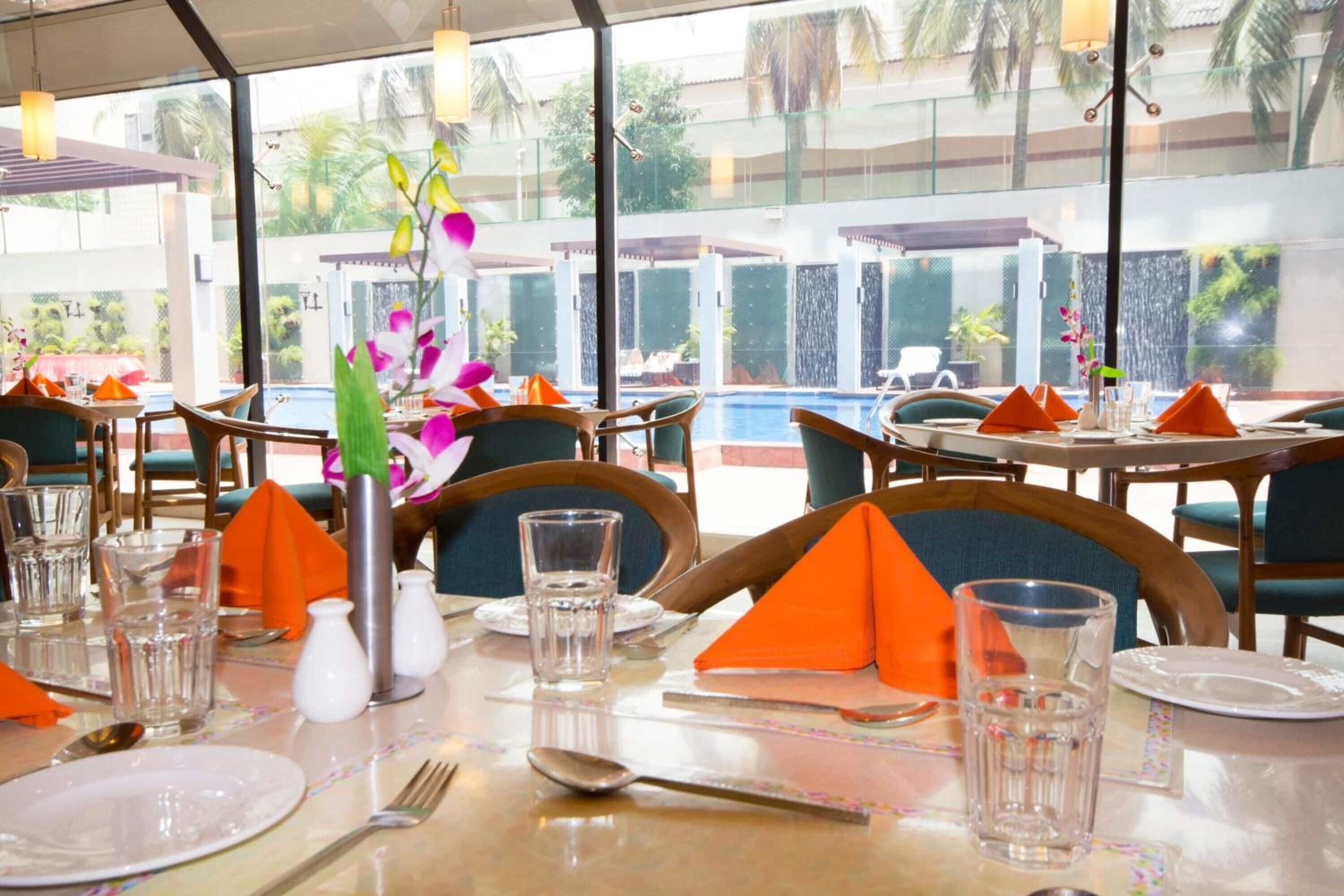 Un restaurant où les tables sont dressées pour le service, avec des serviettes orange, des verres transparents et une orchidée décorative en guise de centre de table.