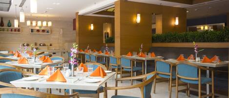 Un restaurante con mesas dispuestas, servilletas anaranjadas, sillas azules y centros de mesa florales.