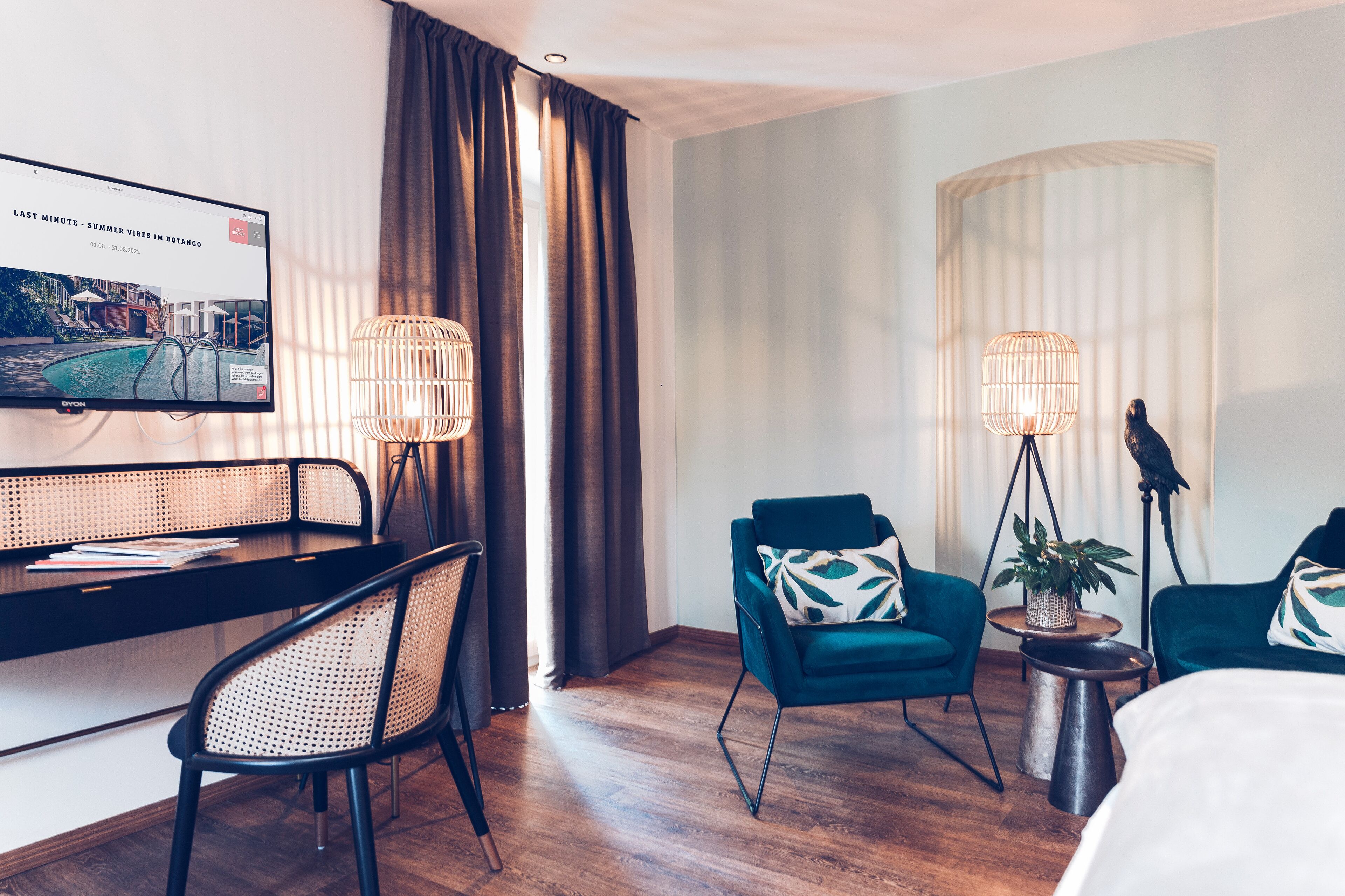 Une chambre d’hôtel avec un plancher en bois, un bureau avec une chaise, deux fauteuils turquoise, un lit, un téléviseur fixé au mur et deux lampadaires.