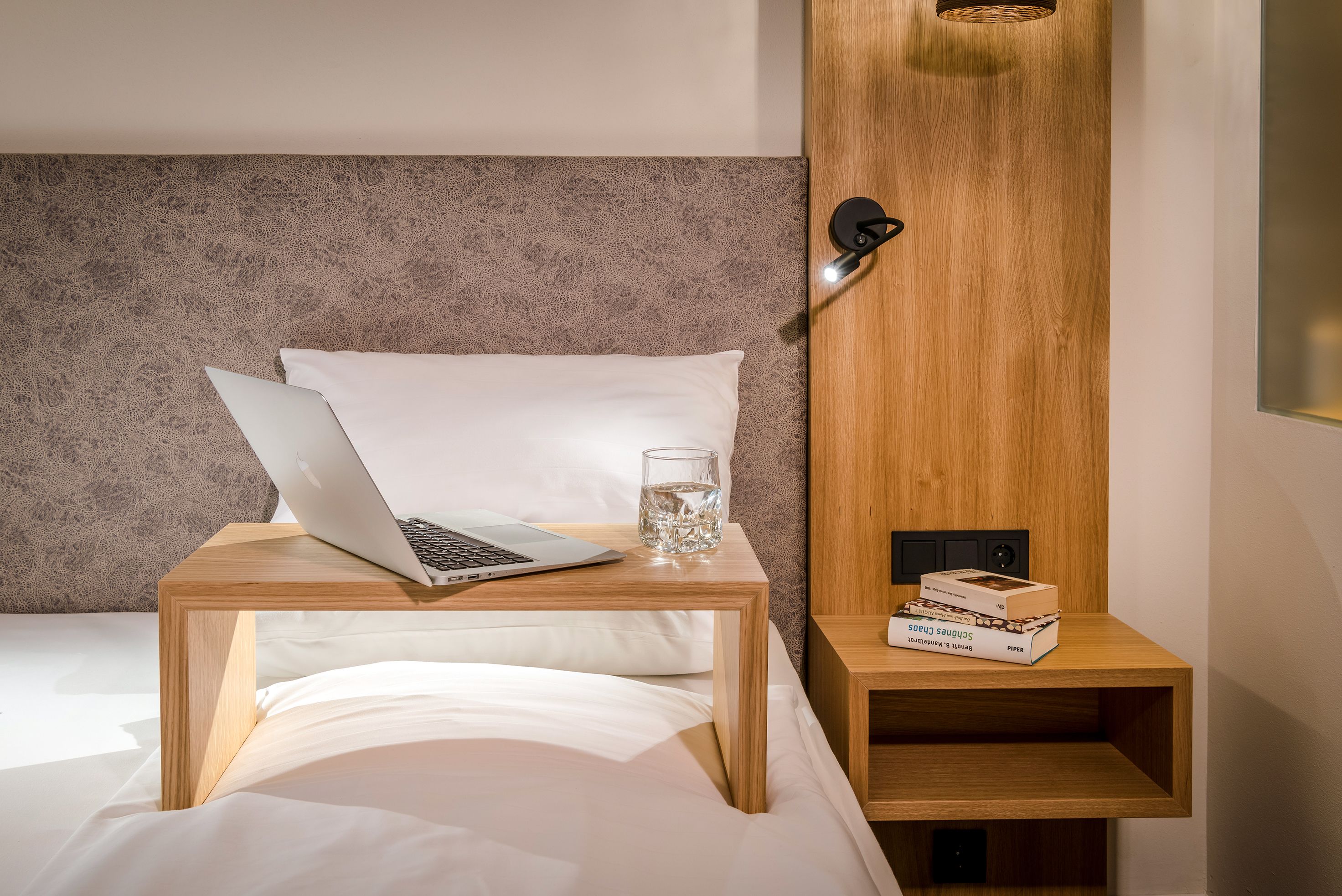 A neatly made bed with white linens, a laptop on a wooden bedside table, a glass of water, and a small wooden shelf with a box and a remote control.