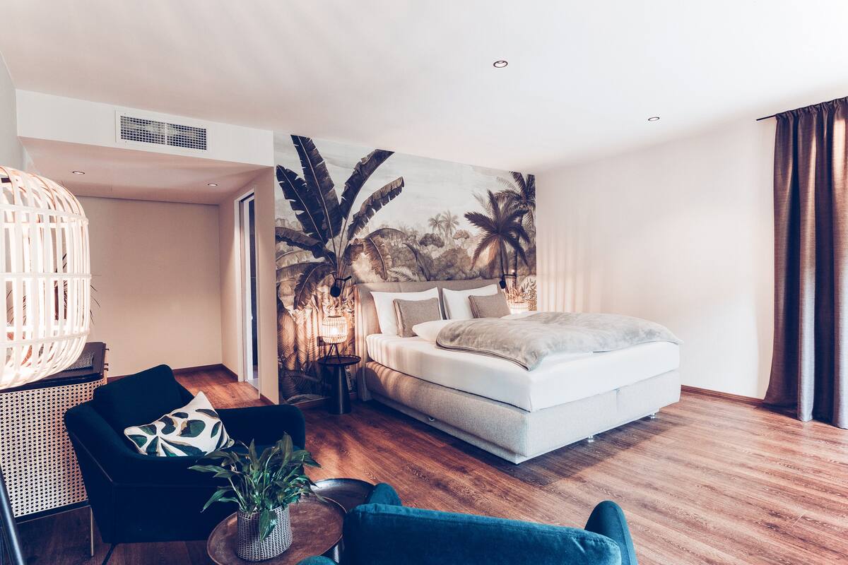 A modern bedroom with a bed, a chair, a small table with a plant, and a wall mural.