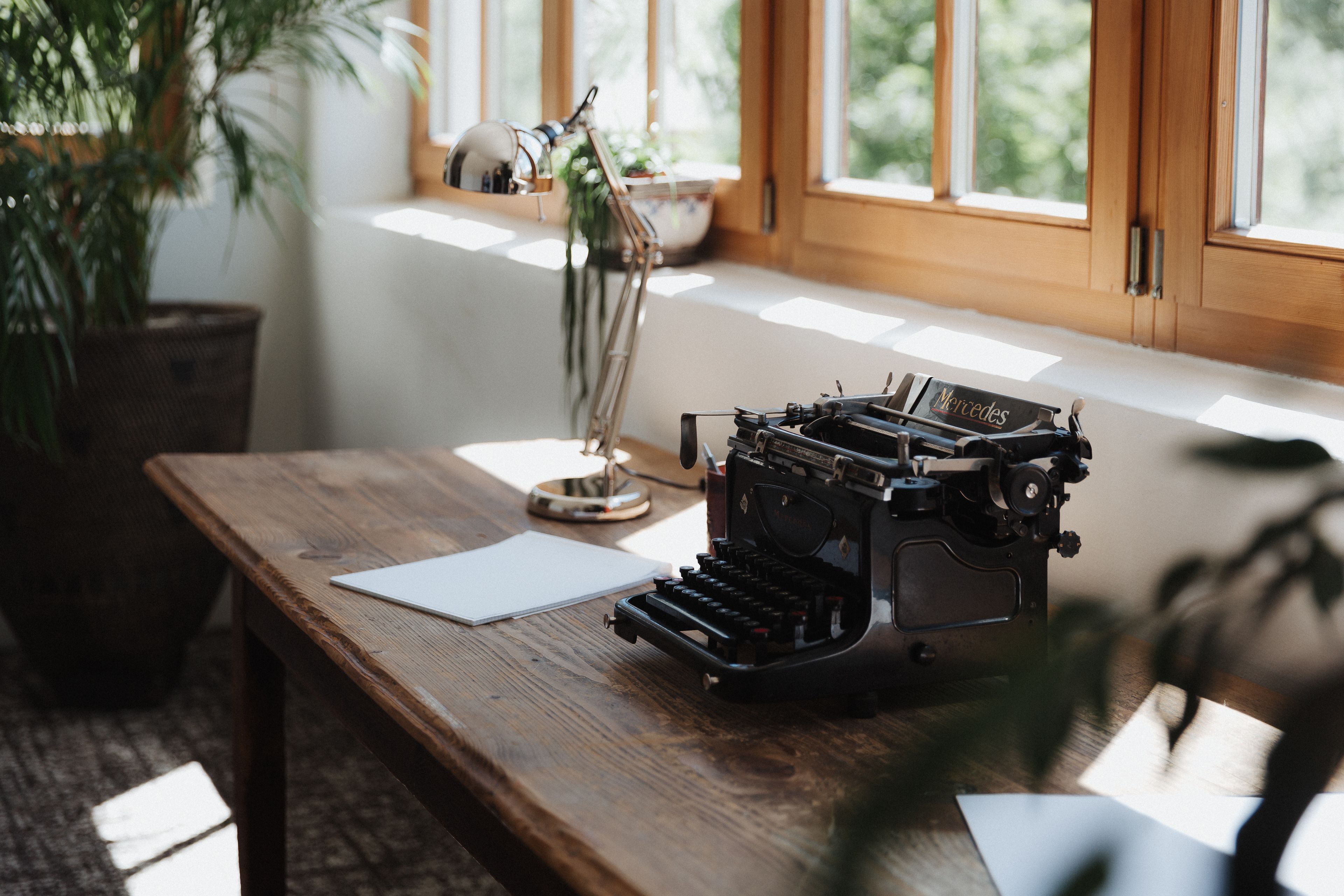 Een houten bureau met een oude schrijfmachine, een potplant en een raam met natuurlijk licht.
