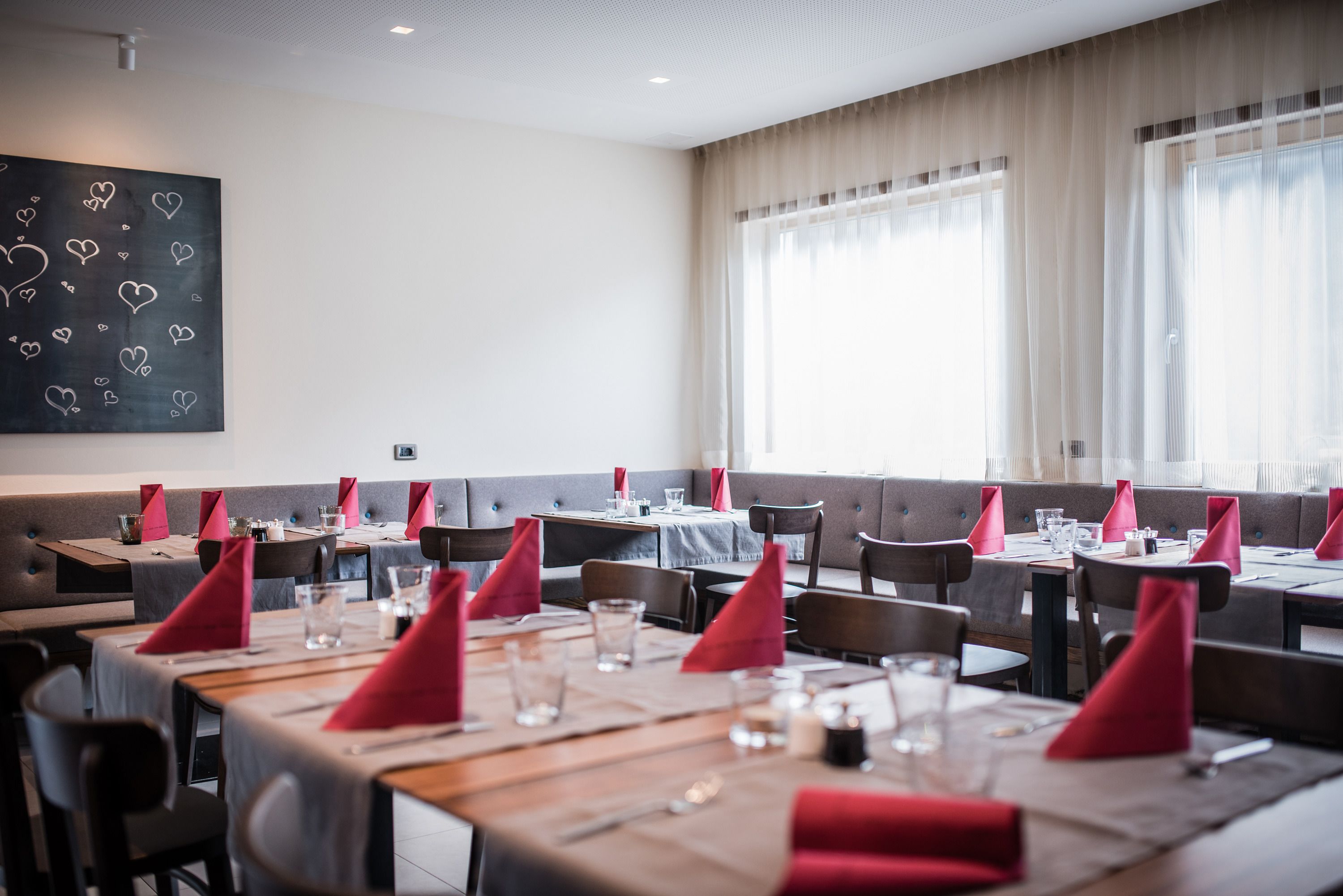 Un restaurante con varias mesas, cada una con servilletas rojas, vasos y cubiertos. Hay cabinas acolchadas a lo largo de las paredes y grandes ventanas con cortinas translúcidas.