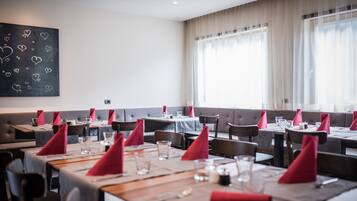 Un restaurante con varias mesas, cada una con servilletas rojas, vasos y cubiertos. Hay cabinas acolchadas a lo largo de las paredes y grandes ventanas con cortinas translúcidas.