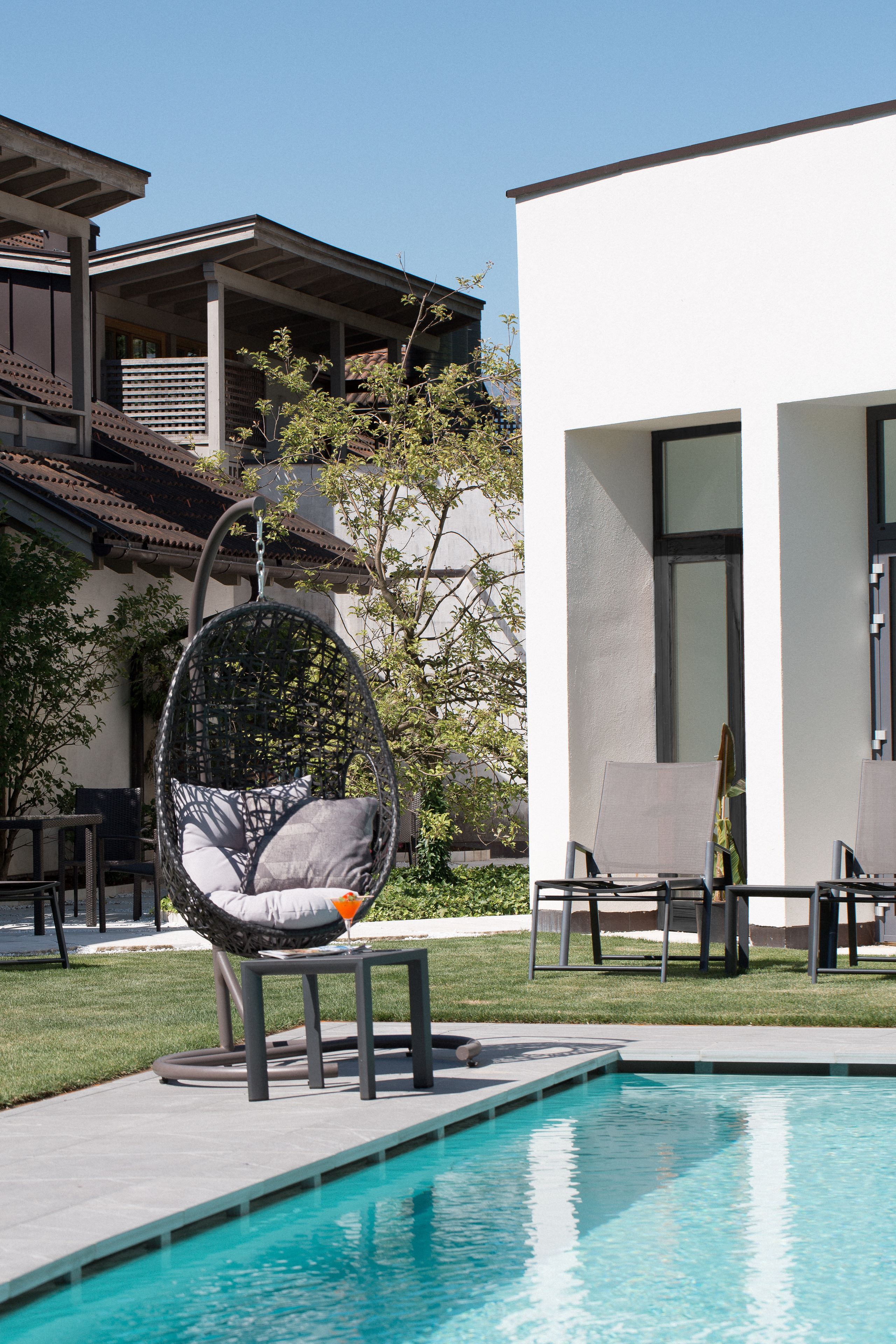 A modern outdoor pool area with a hanging chair, lounge chairs, and a building with large windows.