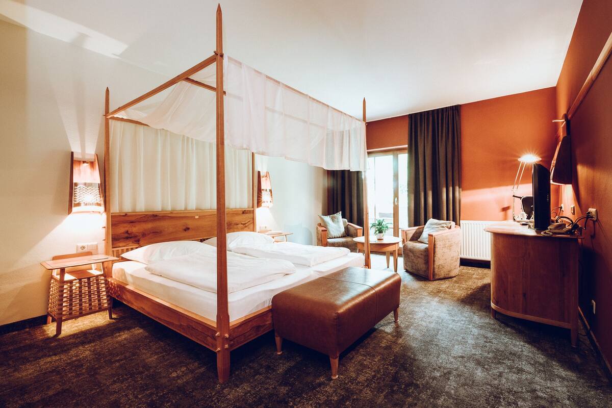 A four-poster bed with a wooden frame and white bedding, a small wooden side table, a brown leather ottoman, a wooden nightstand, a lamp, a chair, and a desk with a computer.