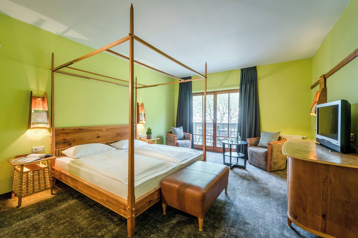 A hotel room with a four-poster bed, a wooden bench, a television, a small table, and a view of the outdoors through large windows.