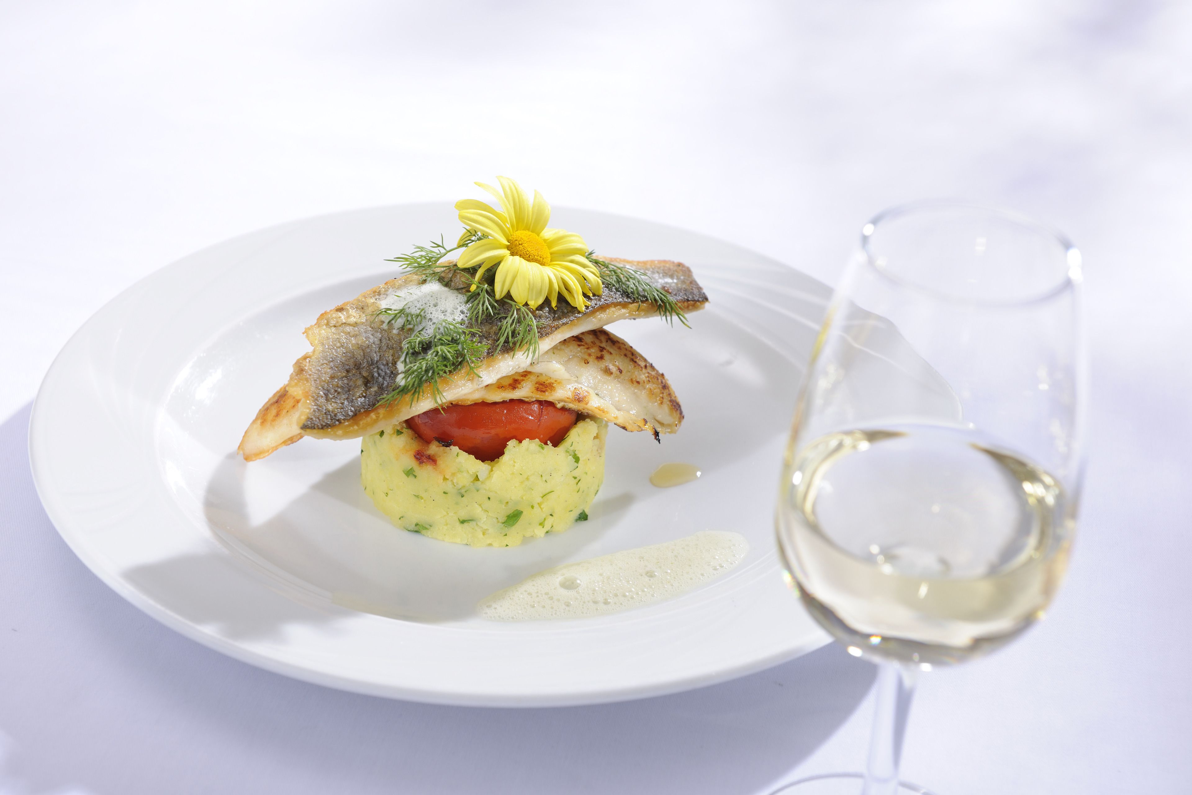 Un plato blanco con un plato de pescado, una copa de vino blanco y un vaso pequeño.