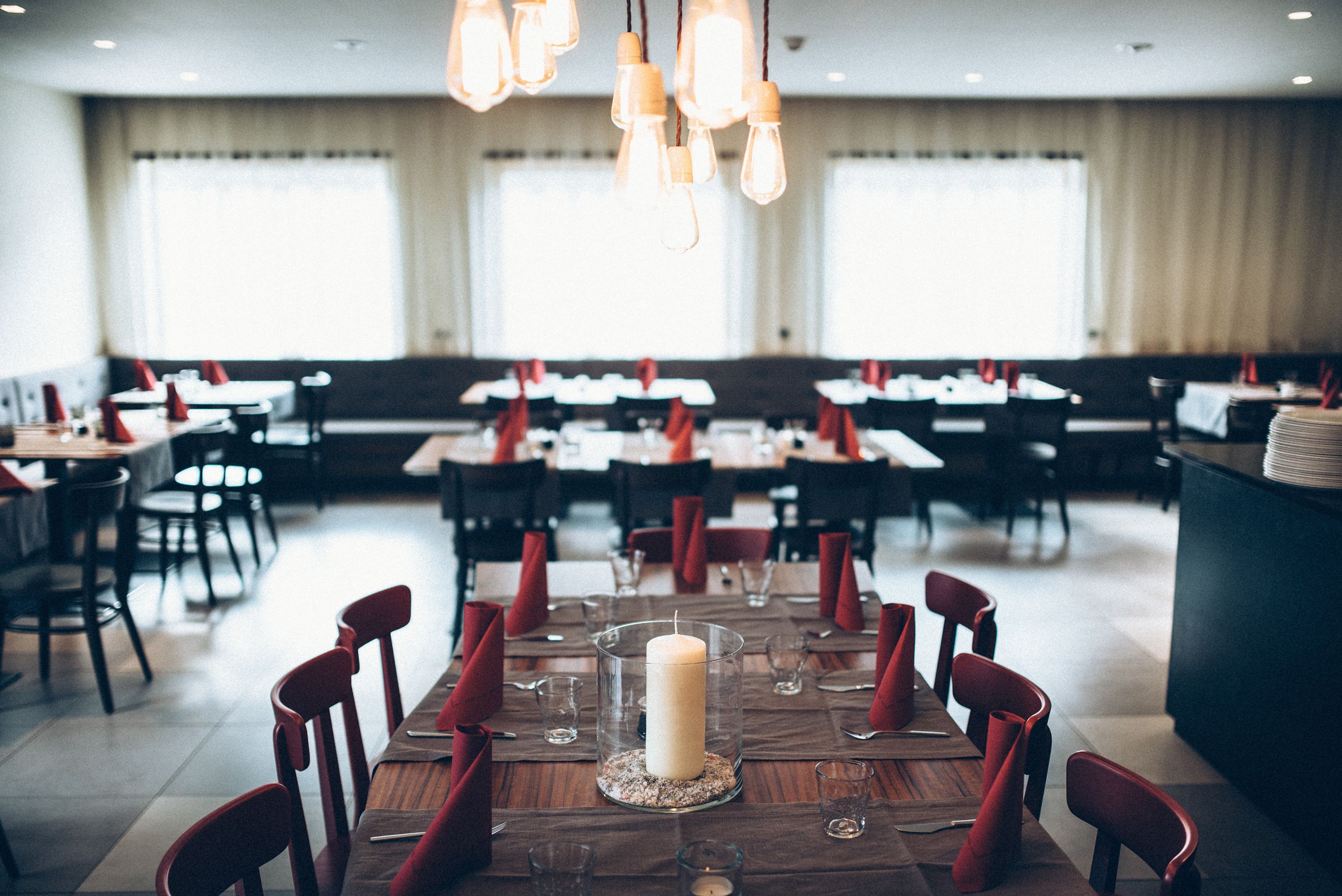 A restaurant with multiple tables set for dining, featuring a central candle centerpiece and red napkins.