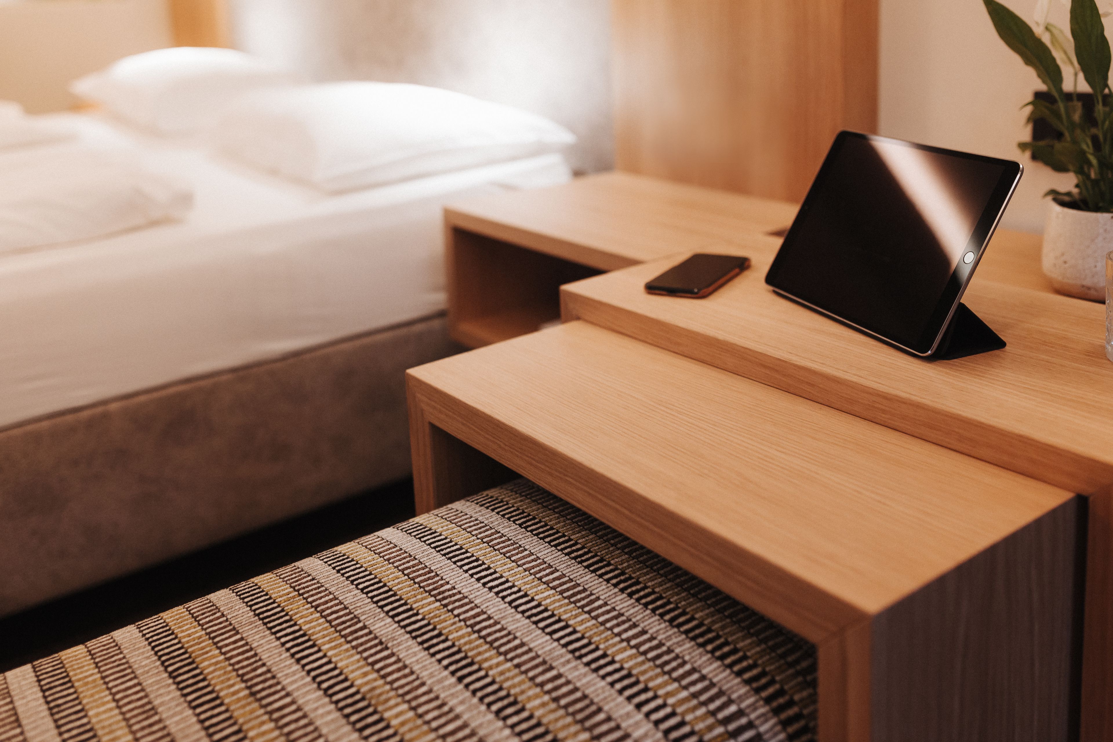 A neatly made bed with white linens, a bedside table with a smartphone and a tablet, and a small potted plant on the nightstand.
