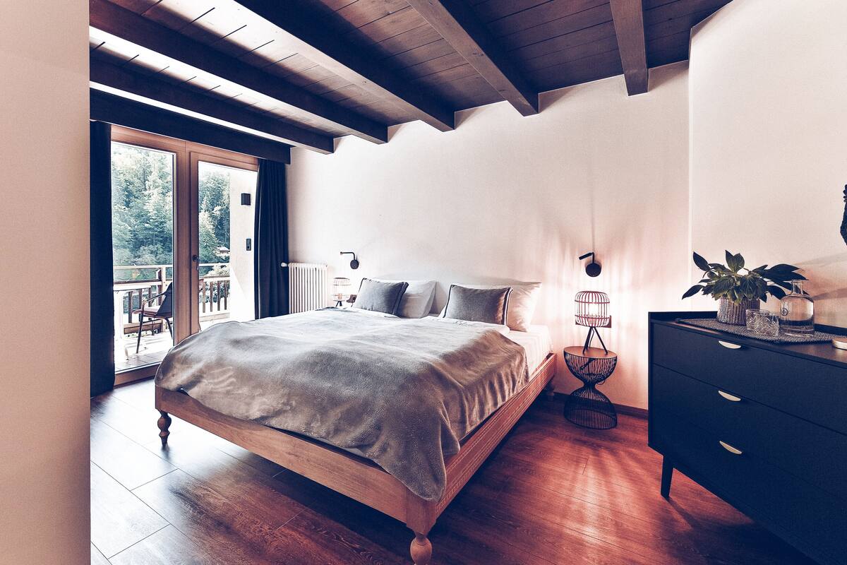 A bedroom with a wooden bed, a black dresser, a small table with a plant, and a view of the outdoors through a sliding glass door.
