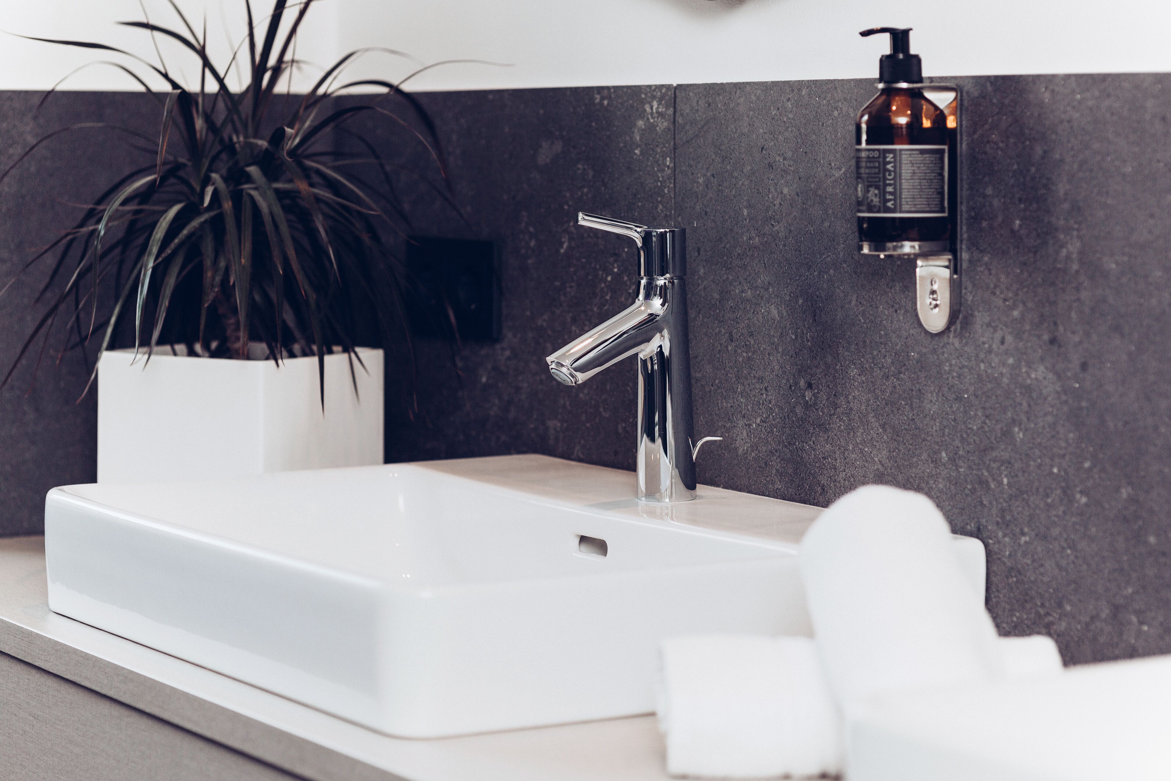 A modern bathroom with a white sink, chrome faucet, and a bottle of shampoo on the wall.
