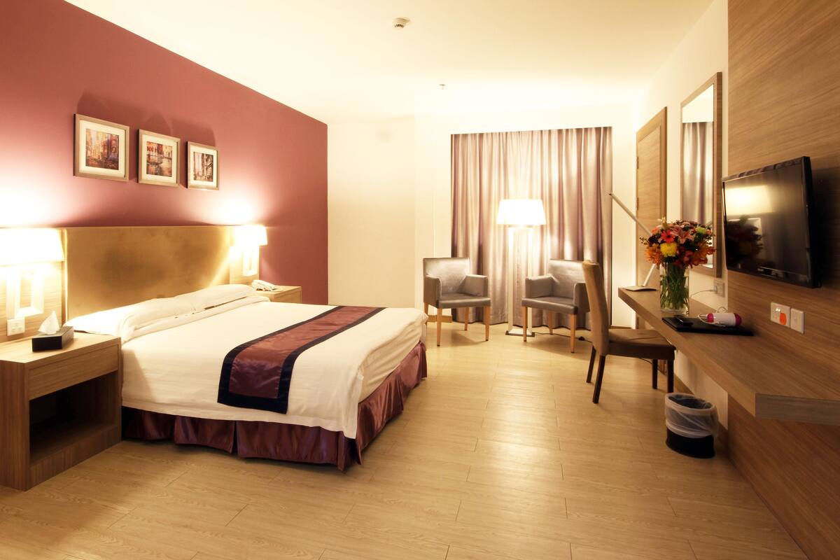 A hotel room with a large bed, bedside tables, a desk with a lamp, two chairs, a TV, and a trash bin.