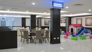 A hotel lobby with a dining area, a children's play area, and a television mounted on a pillar. - Badi'ah Hotel (Bandar Seri Begawan)
