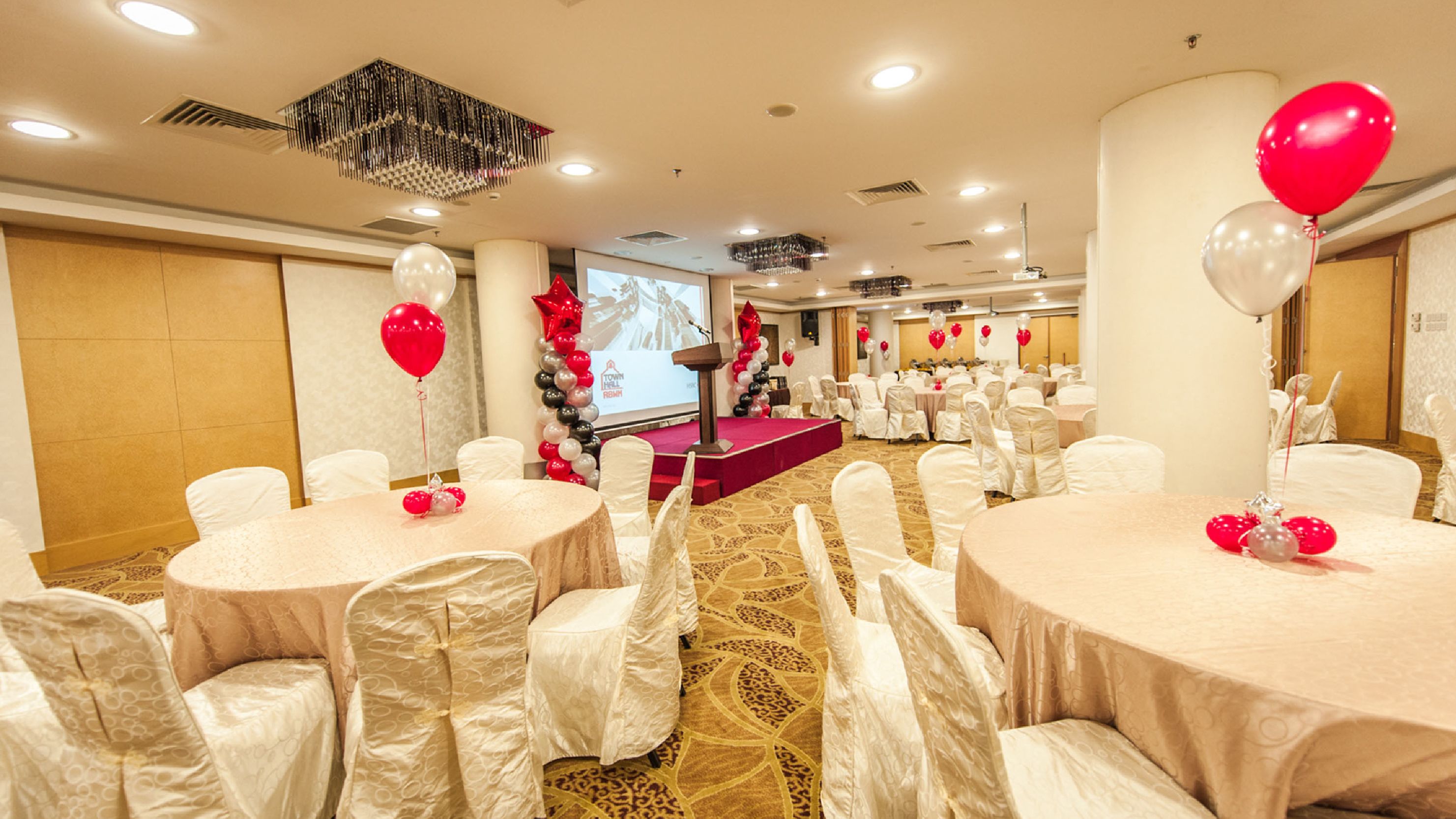 A banquet hall with round tables covered in white tablecloths, white chairs, and red and white balloons.