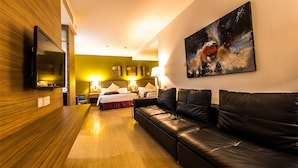 A hotel room with a wooden floor, a black leather sofa, a bed with white linens, two bedside tables with lamps, and a large abstract painting on the wall. - Badi'ah Hotel (Bandar Seri Begawan)
