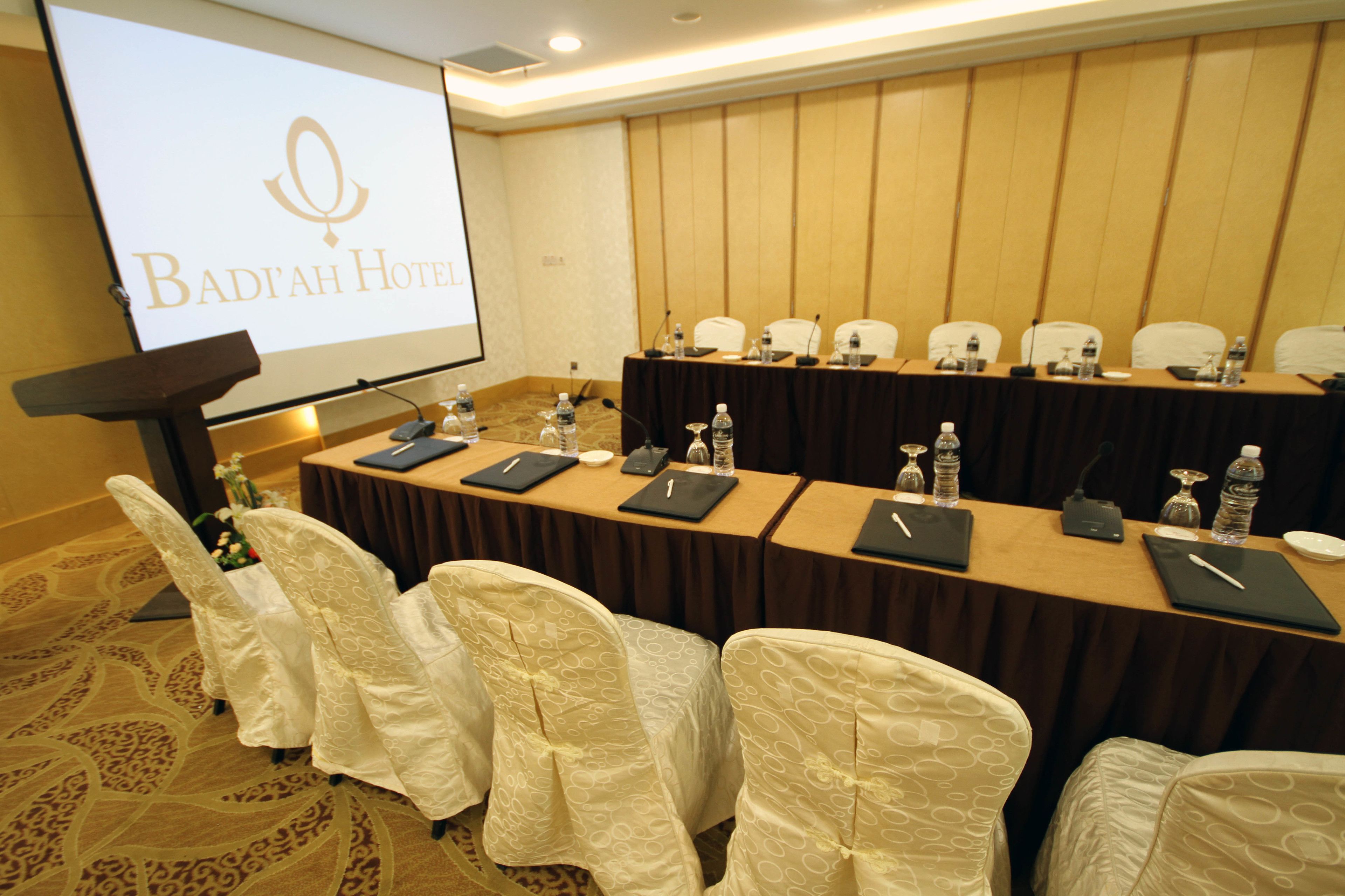 A conference room with a long table, chairs, and a podium with a screen displaying the hotel name.
