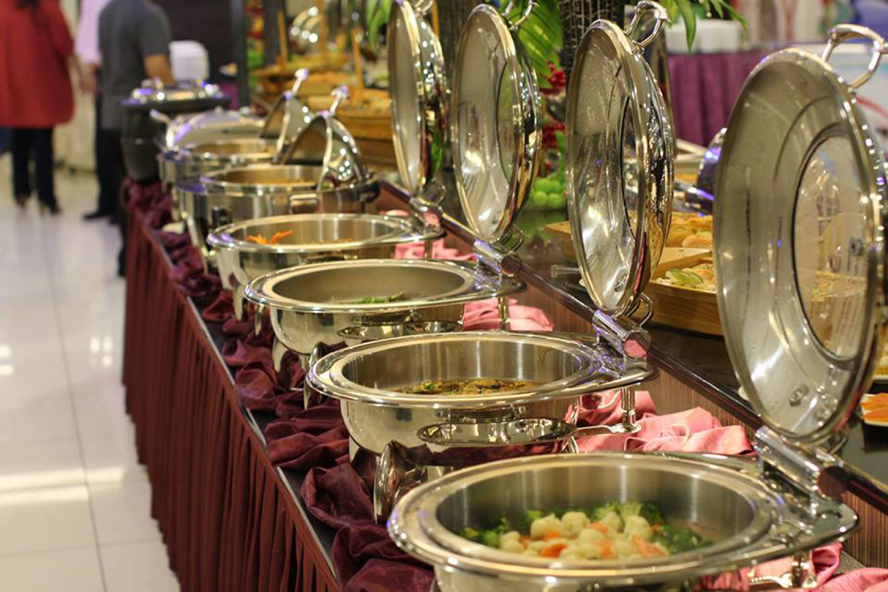 A buffet line with multiple stainless steel serving dishes, some with lids open, revealing food inside.