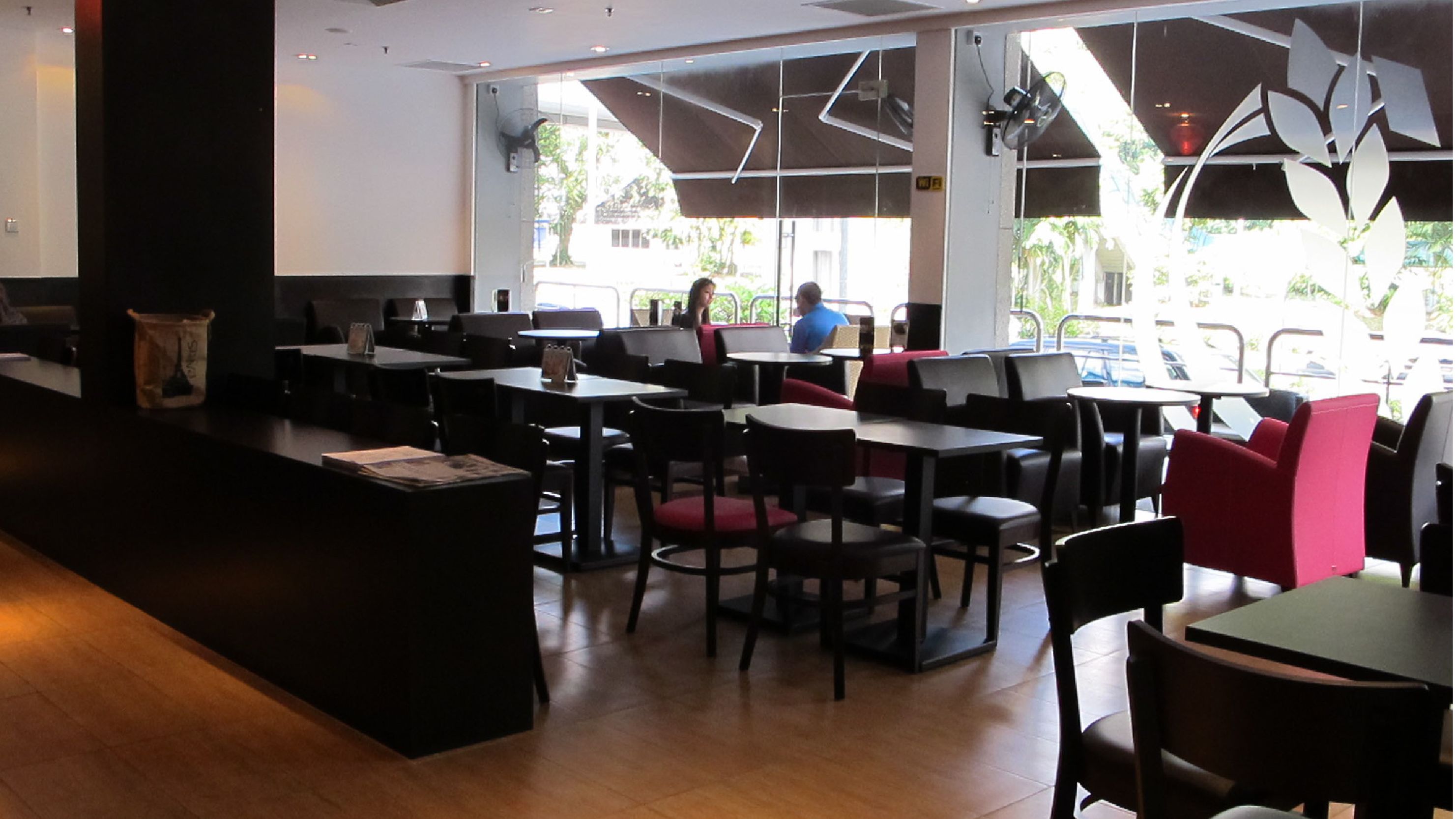 Restaurante com mesas internas, balcão e grandes janelas que permitem entrada de luz natural.