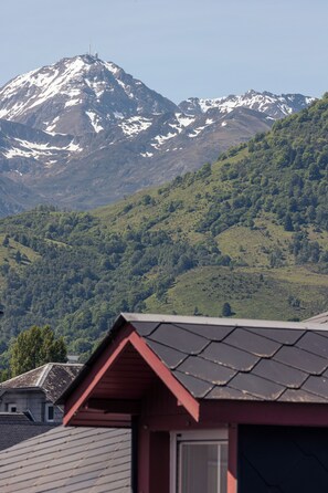 A mountain with snow-capped peaks in the background, a red-roofed building in the foreground, and a forested hillside. - Le Clos du 5 - Appart Hotel Bagneres (Bagnères-de-Bigorre)