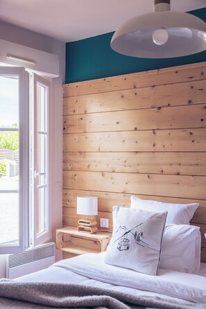 A bedroom with a wooden headboard, a bedside table with a lamp, and a window with a view of greenery. - Le Clos du 5 - Appart Hotel Bagneres (Bagnères-de-Bigorre)