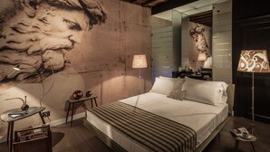 A bedroom with a large mural, a bed with striped bedding, a nightstand, a lamp, and a mirror. - Frattina 57 (Rome)