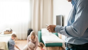 Een man in een blauw shirt die een smartphone gebruikt in een kamer met een groene massagetafel, een spelend kind, en een televisie op de achtergrond.